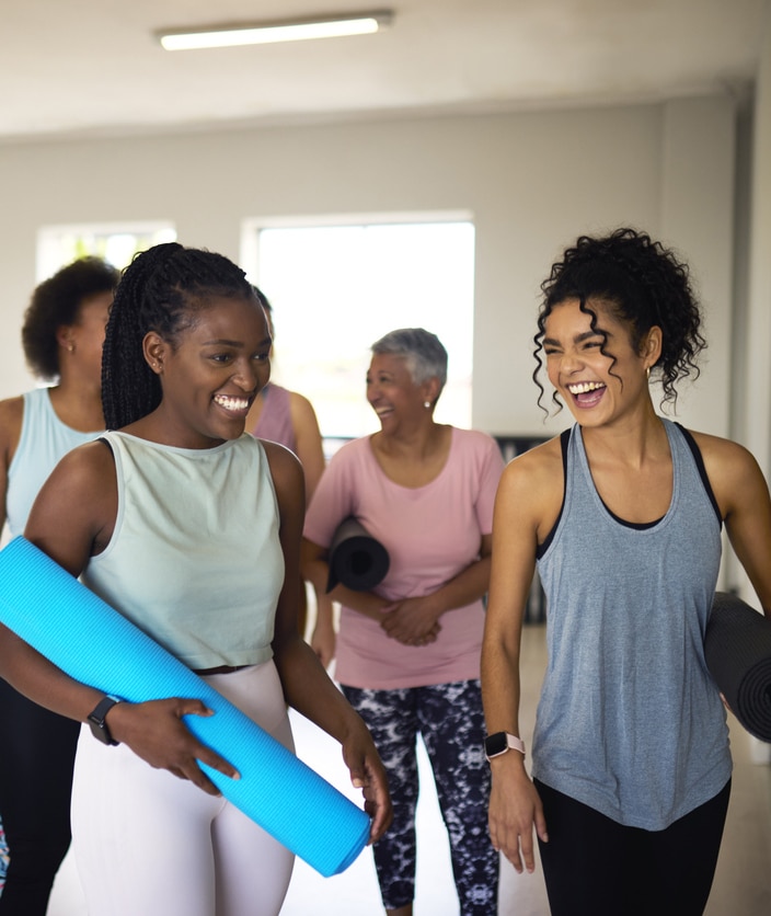 Diverse young student and female coach carrying yoga mats, leaving with friends after yoga class, smiling cheerfully together in studio